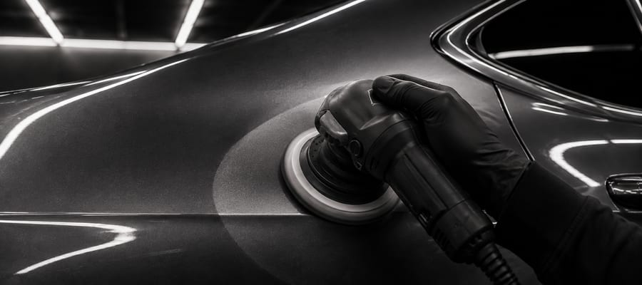 A dual-action polisher working a dark panel under a single light source.